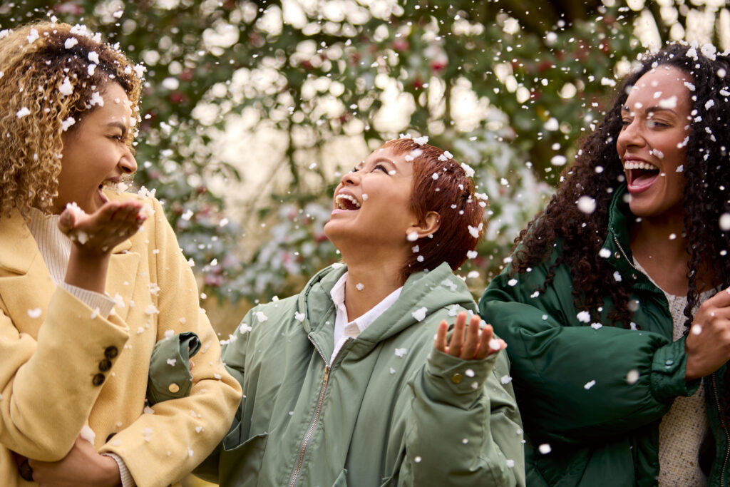three female friends wearing coats standing outside in snowy winter countryside catching snowflakes
